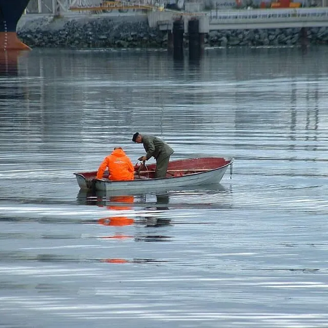 To menn arbeider i liten robåt på stille vann i havnebasseng.