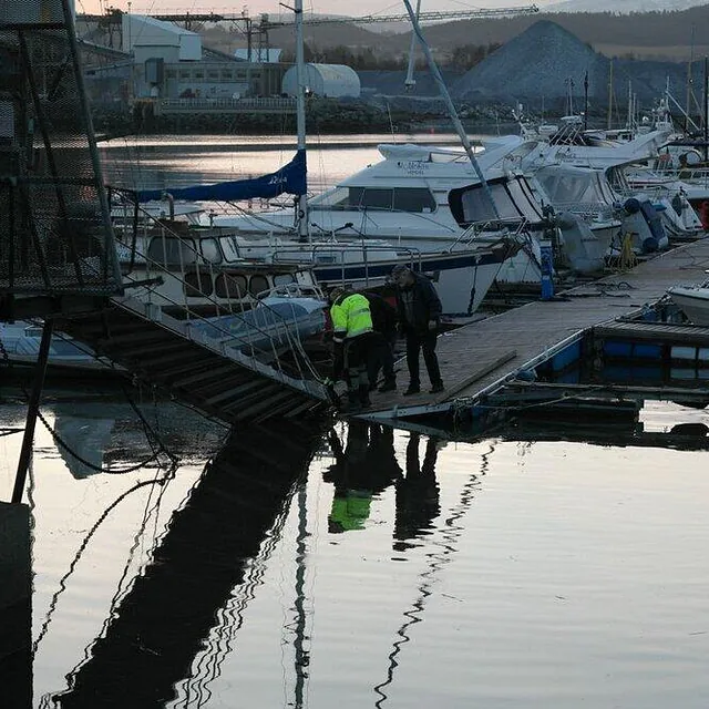 En mann i gul vest jobber nede ved vannet mens to andre ser på fra en brygge i solnedgang.