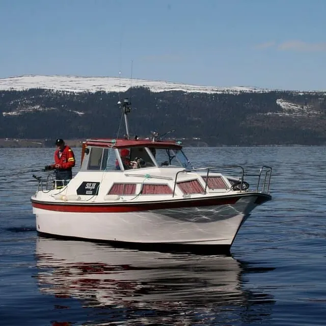 En hvit og rød båt med navnet Silje kjører på sjøen med snødekket fjell i bakgrunnen.