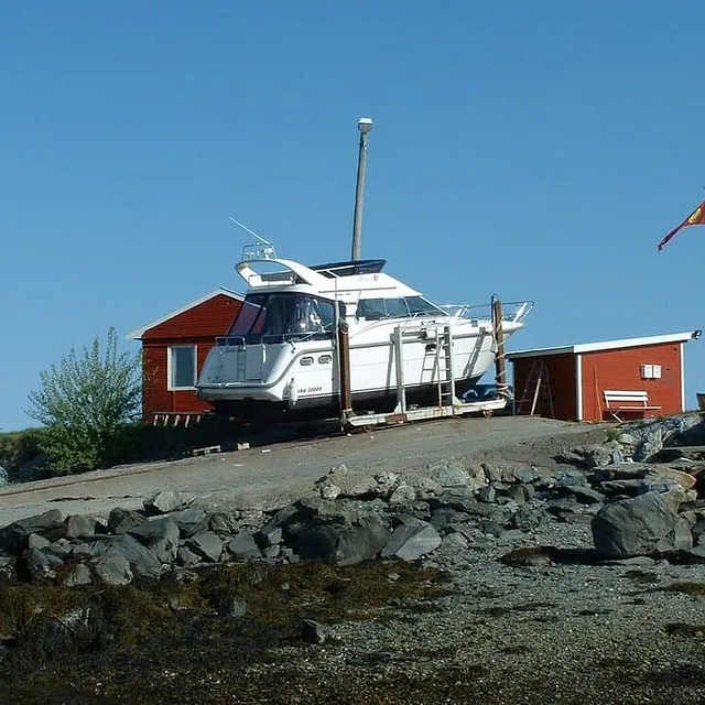 Motorbåt står på land ved røde naust og flaggstang.