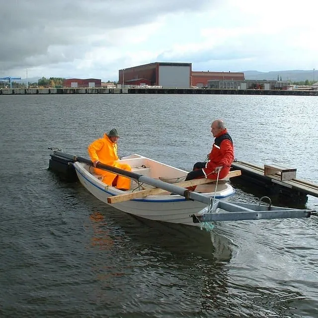 To menn sitter i liten båt ved flytebrygge i industrihavn.