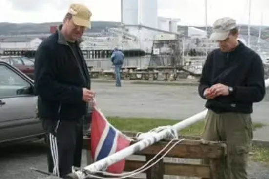 To menn står ved flaggstang og holder norsk flagg i industriområde ved kai.