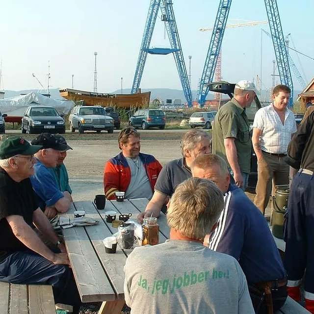 Flere menn samlet rundt piknikbord ved båtverft med kraner i bakgrunnen.