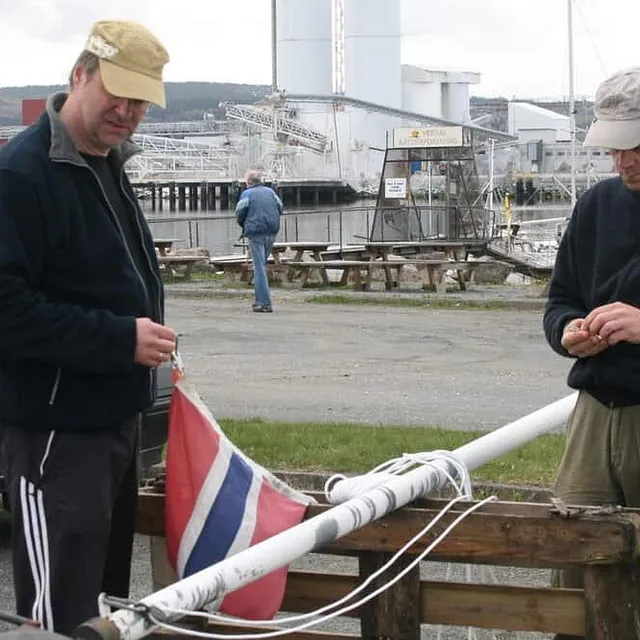 To menn klargjør flaggstang med norsk flagg ved småbåthavn og industriområde i bakgrunnen.