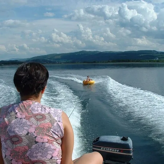 Person sitter bak i båt som trekker oppblåsbar tube over rolig fjord.