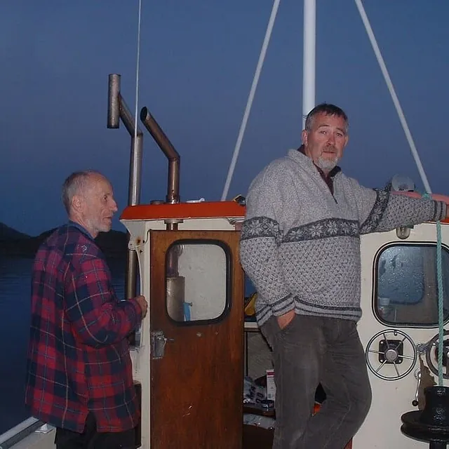 To menn står på dekk av mindre båt i skumring.