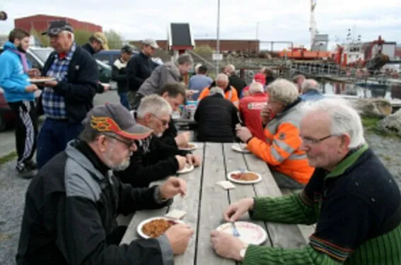 Flere menn sitter ved langbord utendørs og spiser ved kai i industrihavn.