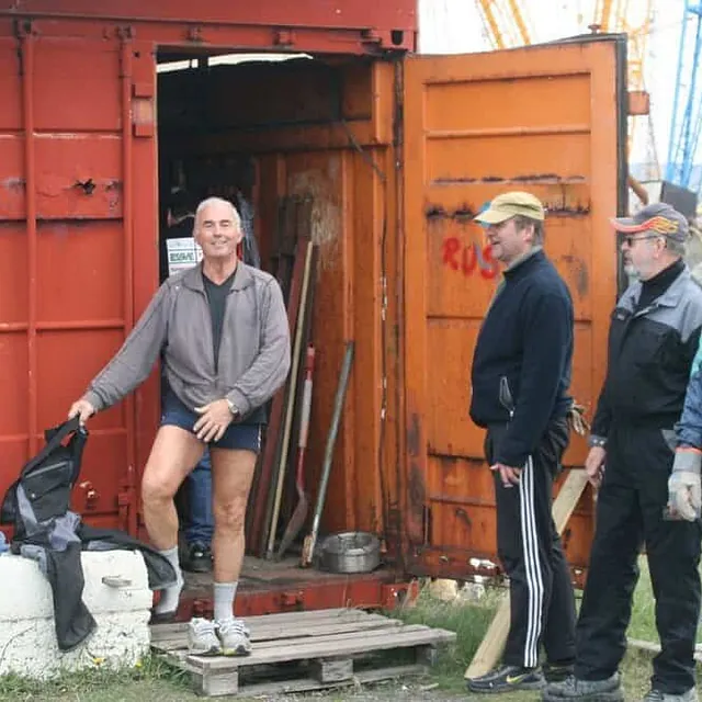Tre menn står utenfor åpen container med verktøy og utstyr ved havneområde.