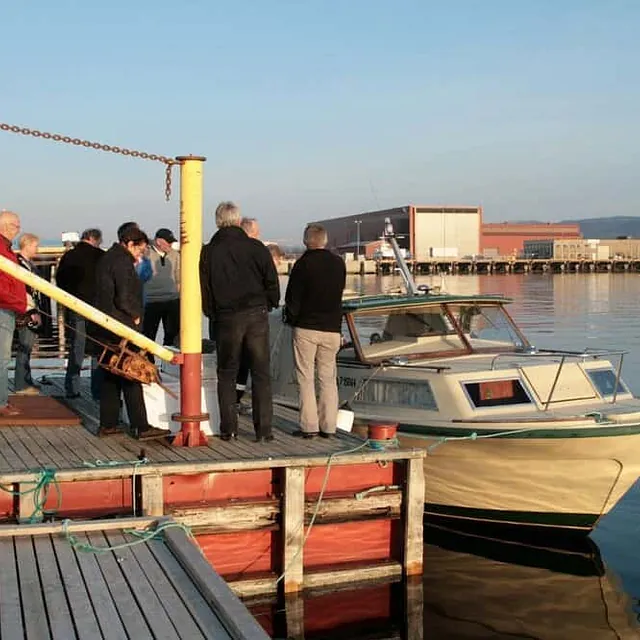 Flere personer står på brygge ved motorbåt i rolig havn.