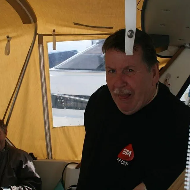 En mann med bart smiler lunt under en gul båtkalesje mens en annen mann sitter bak ham.