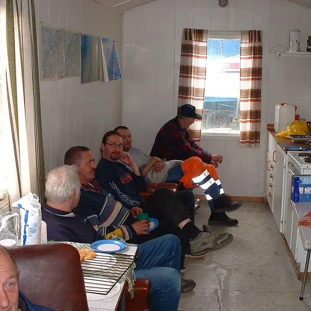 Flere menn sitter samlet i liten brakke med kaffekopper og samtale.