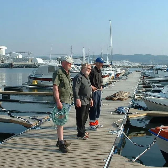 Tre menn står på brygge omgitt av fritidsbåter i solskinn.