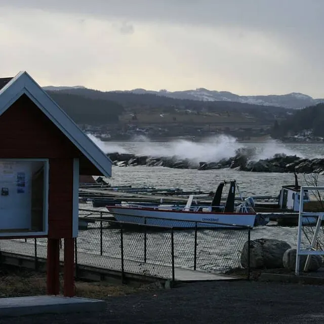 En liten rød bu ved en brygge i en småbåthavn mens bølgene slår mot moloen.