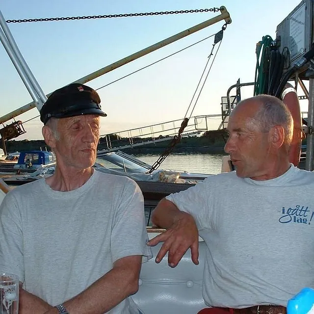 To eldre menn sitter og prater om bord i båt ved kai i sollys.