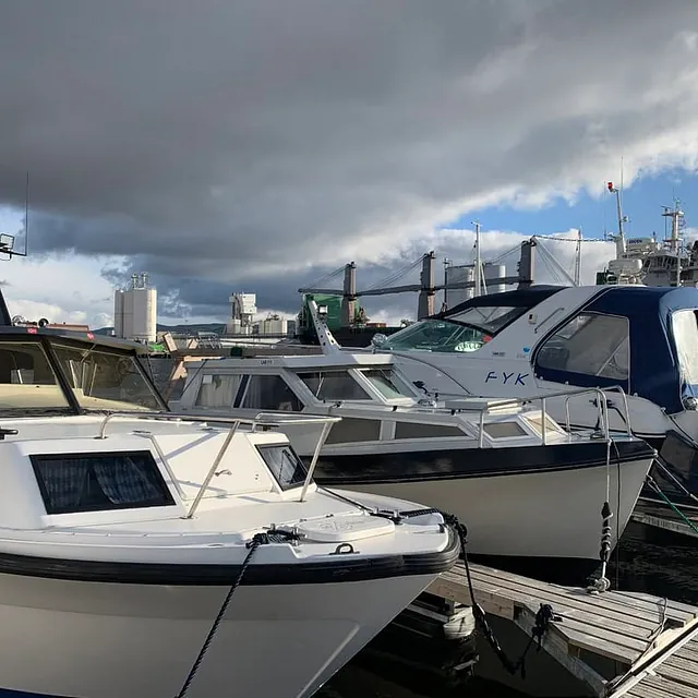 Motorbåter fortøyd ved brygge i industrihavn med kraner og lagerbygg bak.