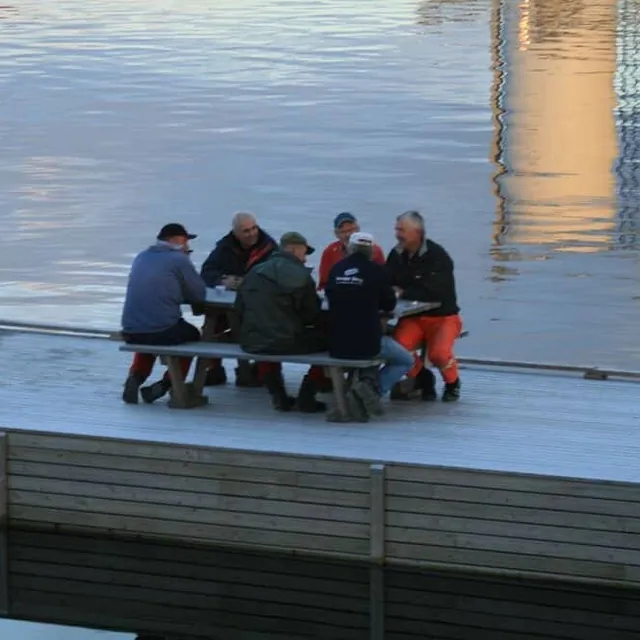Flere menn sitter rundt bord på brygge ved rolig vann.