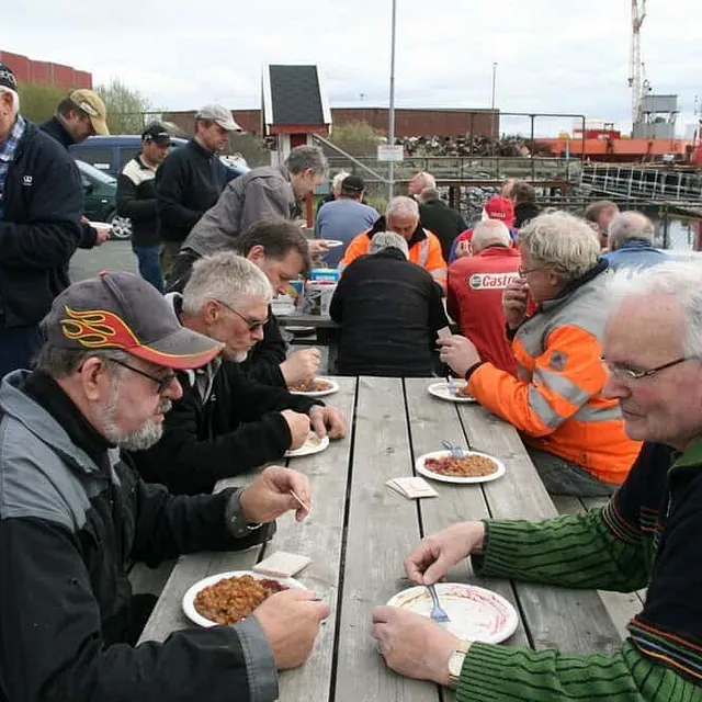 Flere menn sitter ved langbord utendørs og spiser ved havn.
