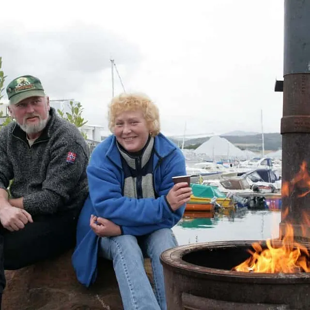 En mann og en dame sitter ute ved en varm bålpanne med en småbåthavn i bakgrunnen.