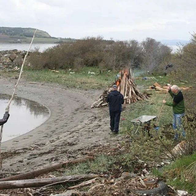 Tre menn rydder drivved og brenner bål på strand ved fjord.