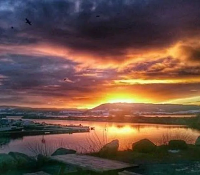 En fargerik og dramatisk solnedgang over en rolig småbåthavn med mørke skyer.