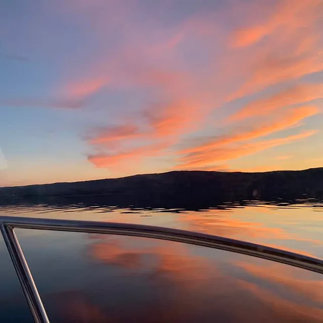 Solnedgang over fjord sett fra båt med rosa skyer og speiling i vannet.