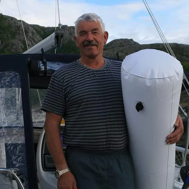 Eldre mann holder stor hvit fender på dekk foran fritidsbåt i havn.