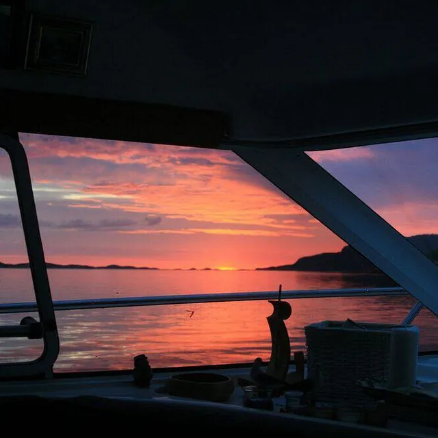 Solnedgang sett fra båtinteriør med utsikt over rolig sjø og øyer.