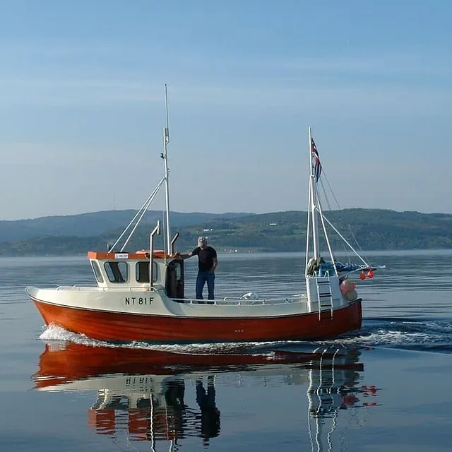 Sjark går sakte over blank fjord med speiling i vannet.