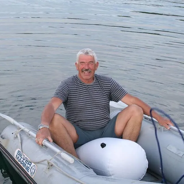 Eldre mann sitter smilende i liten gummibåt på stille sjø.