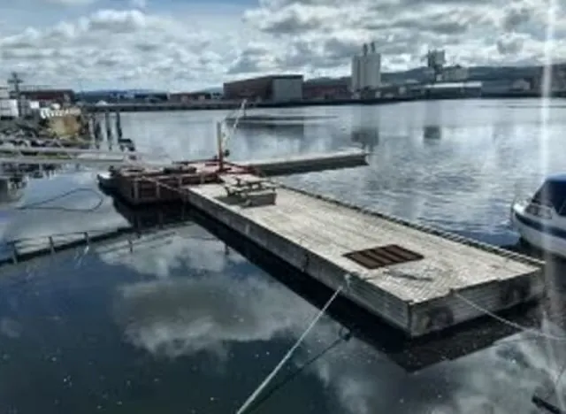 Flytebrygge i industrihavn med stille vann og lagerbygg i bakgrunnen.