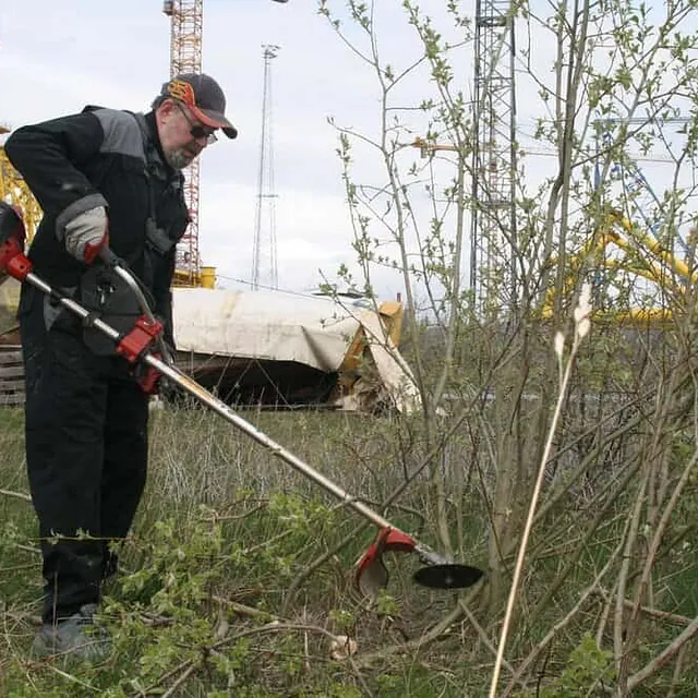 Mann rydder kratt med motorisert ryddesag i industriområde nær havn.