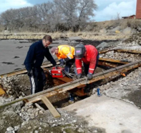 Tre arbeidere reparerer skinnegang med verktøy på gruslagt område ved sjøen.