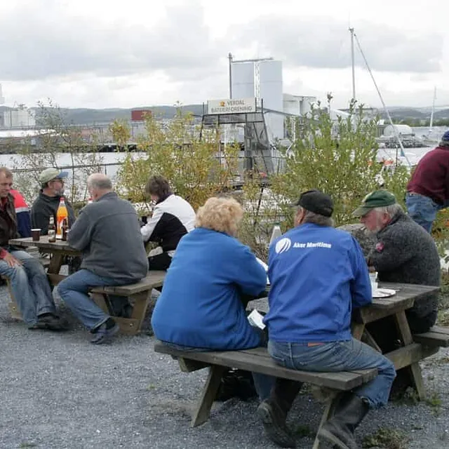 Folk sitter på benker og prater ute ved havna med industribygg og fjell i det fjerne.