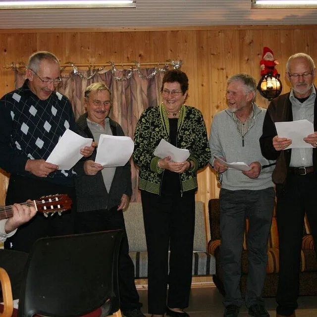 En gruppe glade pensjonister synger sammen fra ark mens en mann spiller akustisk gitar.