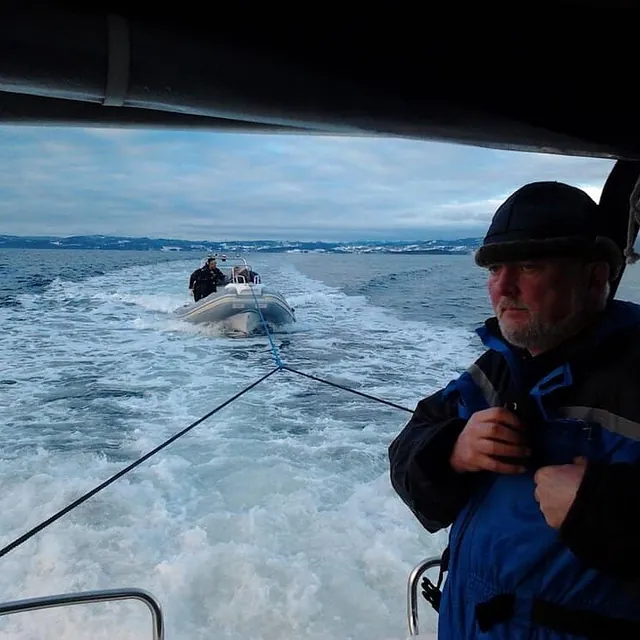 Mann står i båt mens mindre båt slepes bak over bølgende sjø.