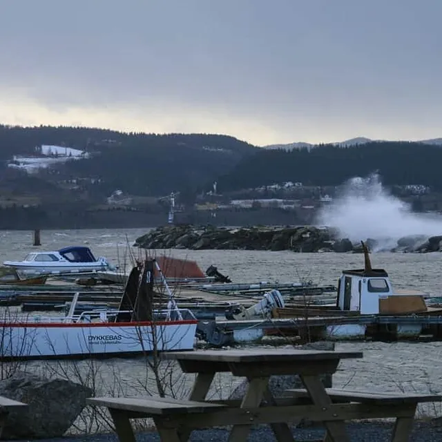 Bølger slår over en molo i en havn med småbåter og skogkledde åser i bakgrunnen.
