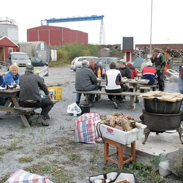 En gruppe mennesker sitter ved langbord ute og spiser krabber fra en stor hvit kasse.