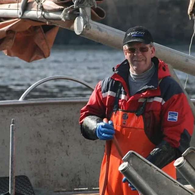 En smilende mann i rød og oransje arbeidsdress og caps står om bord i en båt på sjøen.
