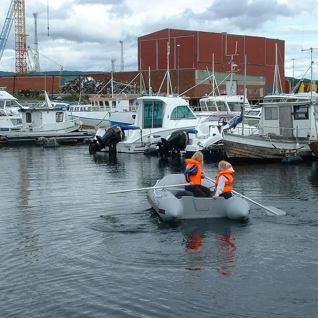 To barn i redningsvester ror liten jolle mellom fortøyde båter i havn.