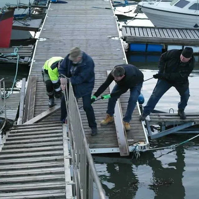 Tre menn bruker muskelkraft og tau for å flytte en metallskinne på en trebrygge ved sjøen.