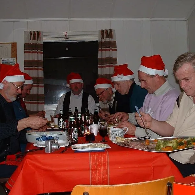 Flere menn med nisseluer sitter samlet rundt bord med julemat innendørs.