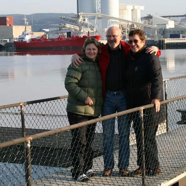 Tre personer står på brygge foran industrihavn med skip og lagerbygg i bakgrunnen.