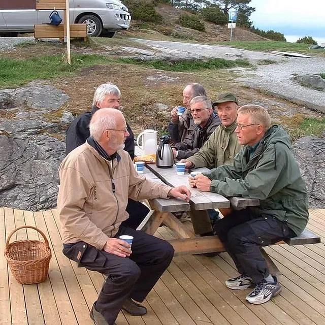 Fem menn sitter rundt piknikbord og drikker kaffe utendørs.