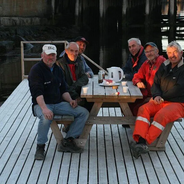 Seks menn sitter ved bord på brygge under kai i kveldssol.