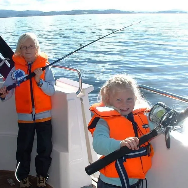 To barn i redningsvester holder fiskestenger om bord i båt.
